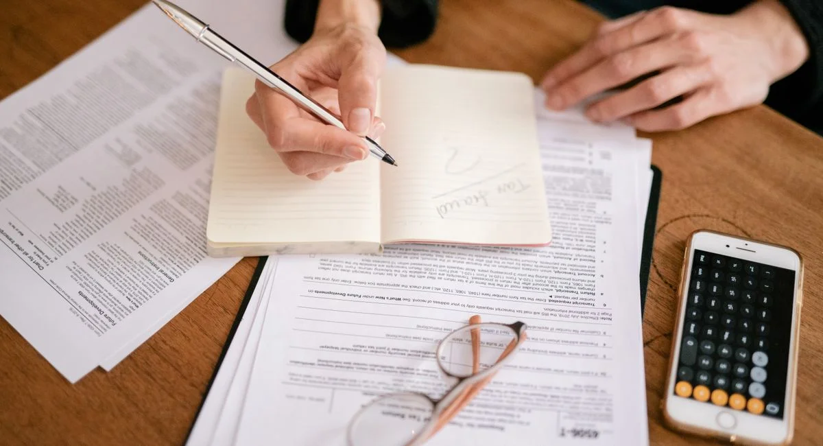 Close-up of a person writing in a notebook with documents and a calculator, managing finances.