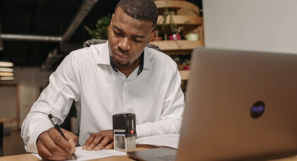 A young man in a white shirt works diligently at his office desk with a laptop and documents.