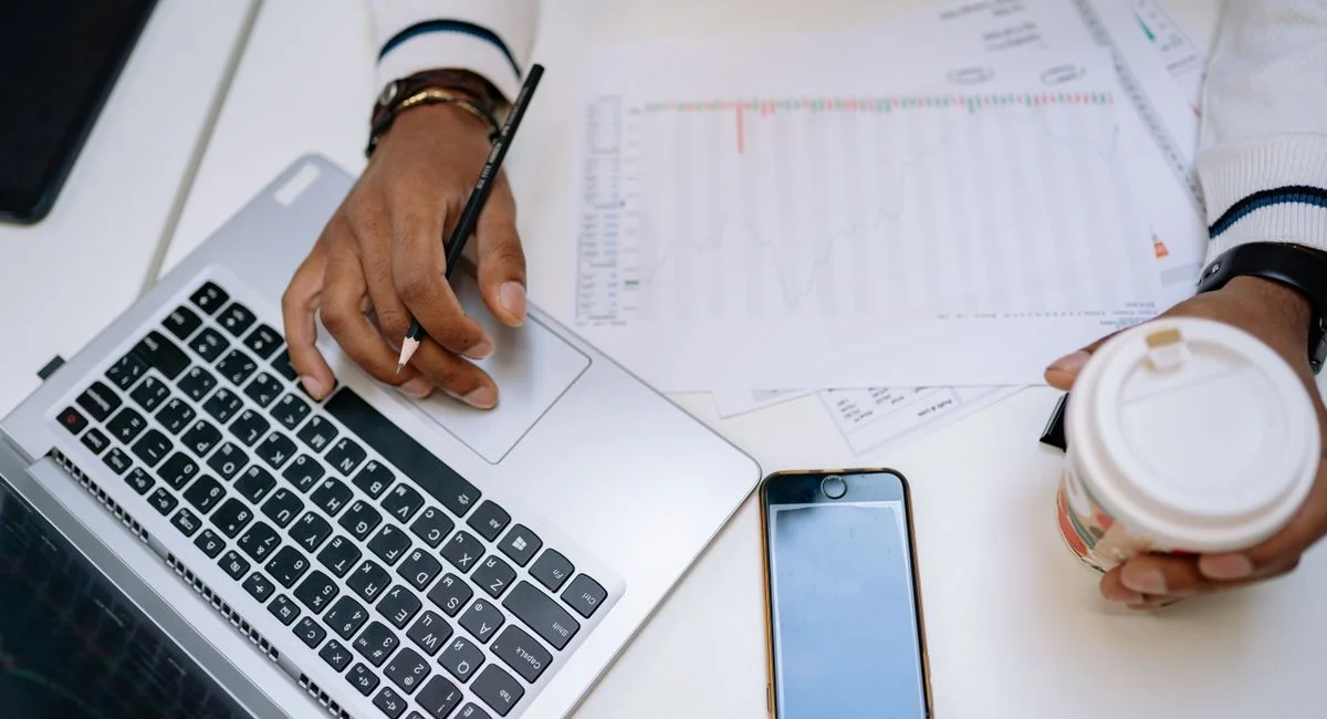 A businessman holding coffee and using a laptop at a desk with charts and a smartphone.