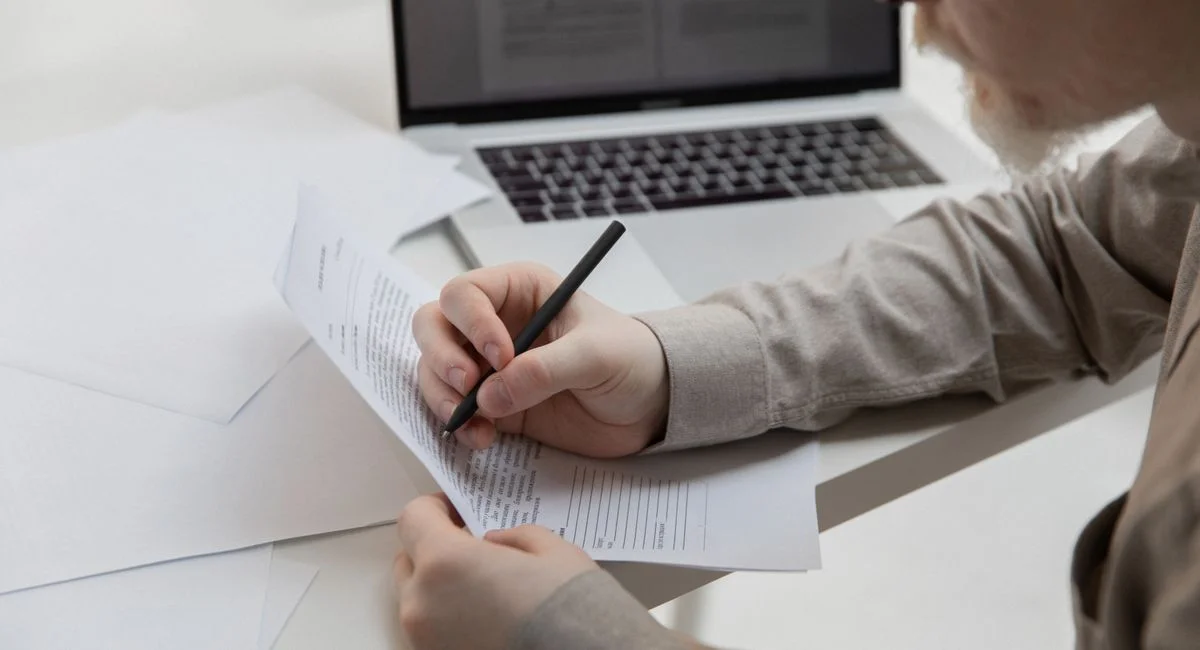 Side view of crop anonymous manager with pen watching paper with text near netbook on desk