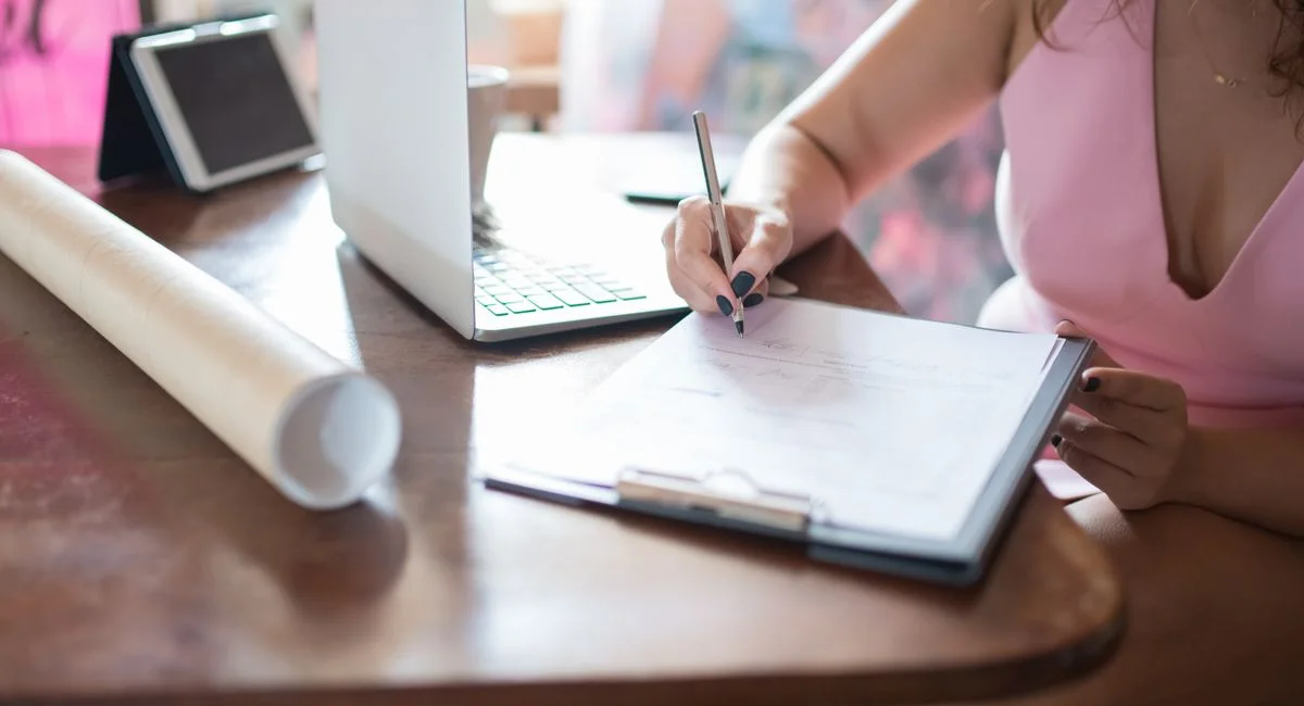 Businesswoman writing on a clipboard at a table with a laptop indoors.