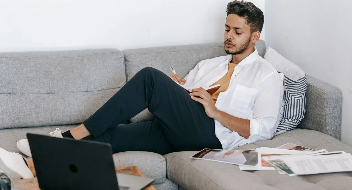 Side view of young ethnic male journalist sitting on sofa and writing ideas for new article in notebook while working with la