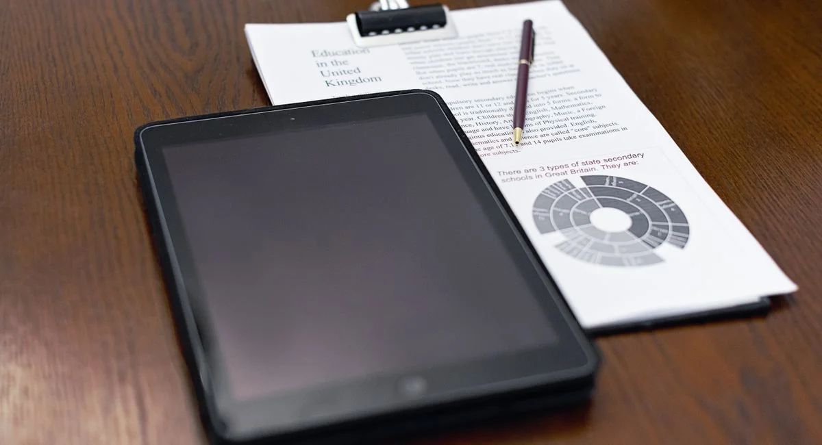 A tablet and educational documents on a wooden table with a pen.