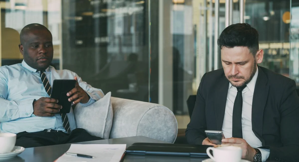 Two businessmen focused on tablets and phones in modern office lounge.