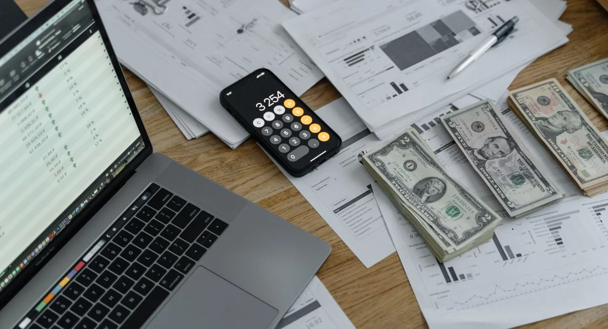 A desk with financial documents, currency, a laptop, and phone calculator.