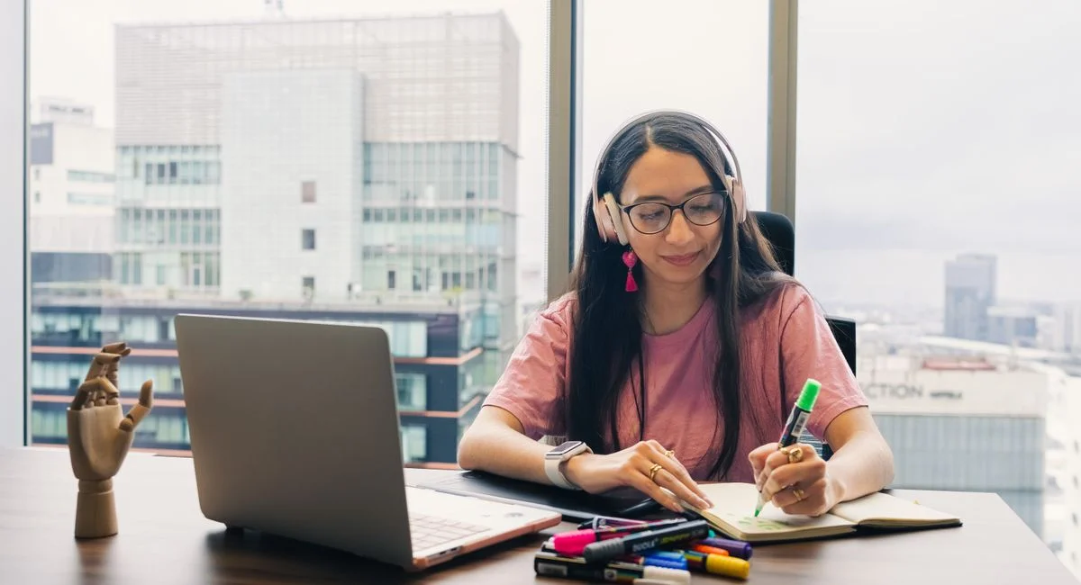 Young woman wearing headphones, writing in notebook at office desk with city view.