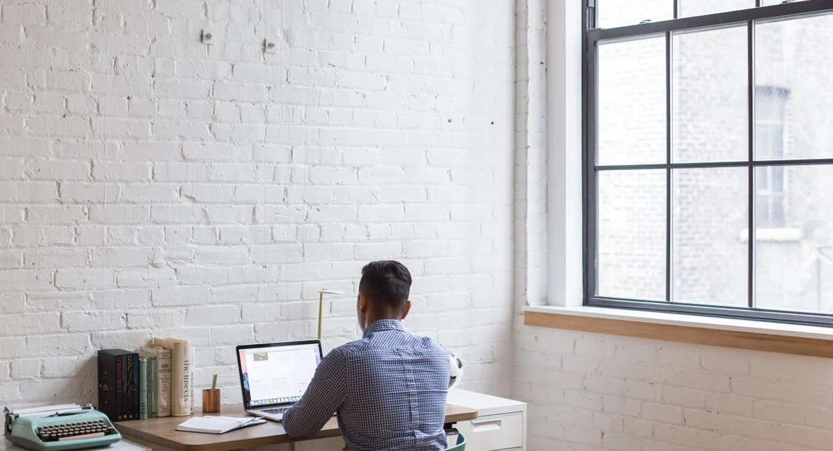 Adult male working remotely on a laptop in a well-lit home office with a large window.