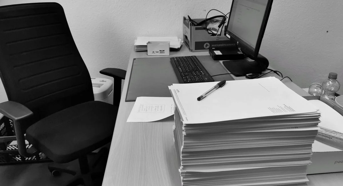 Black and white image of a cluttered office desk with a computer, chair, and paperwork.