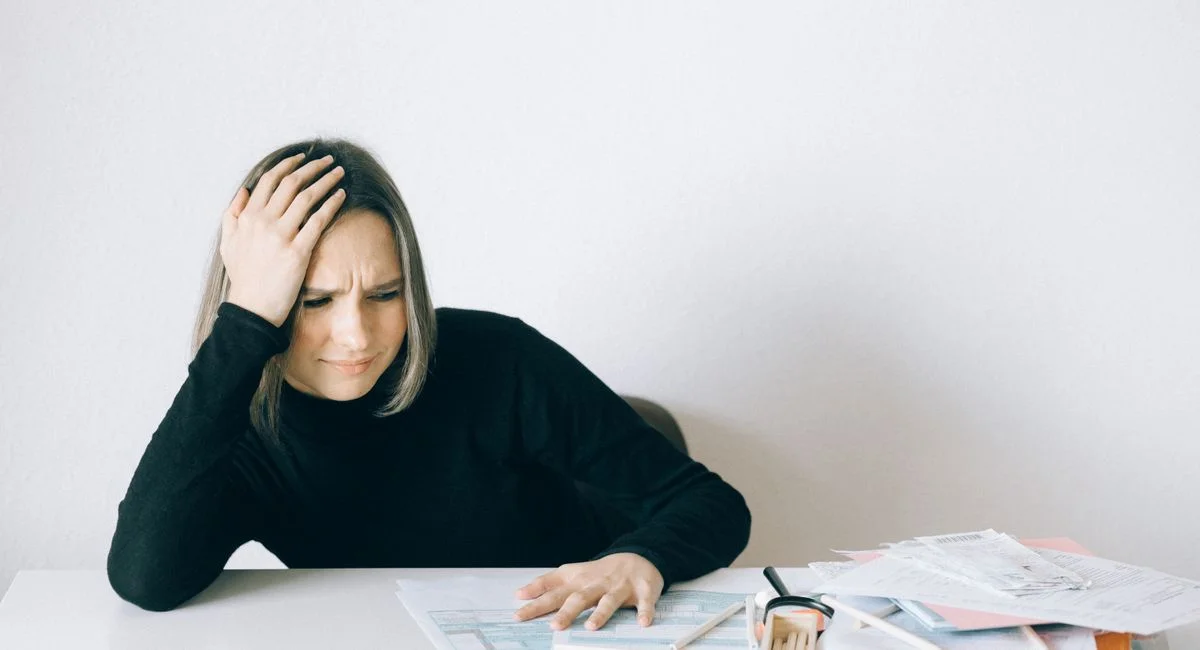 Woman in black sweater stressed with financial paperwork, overwhelmed at white table.