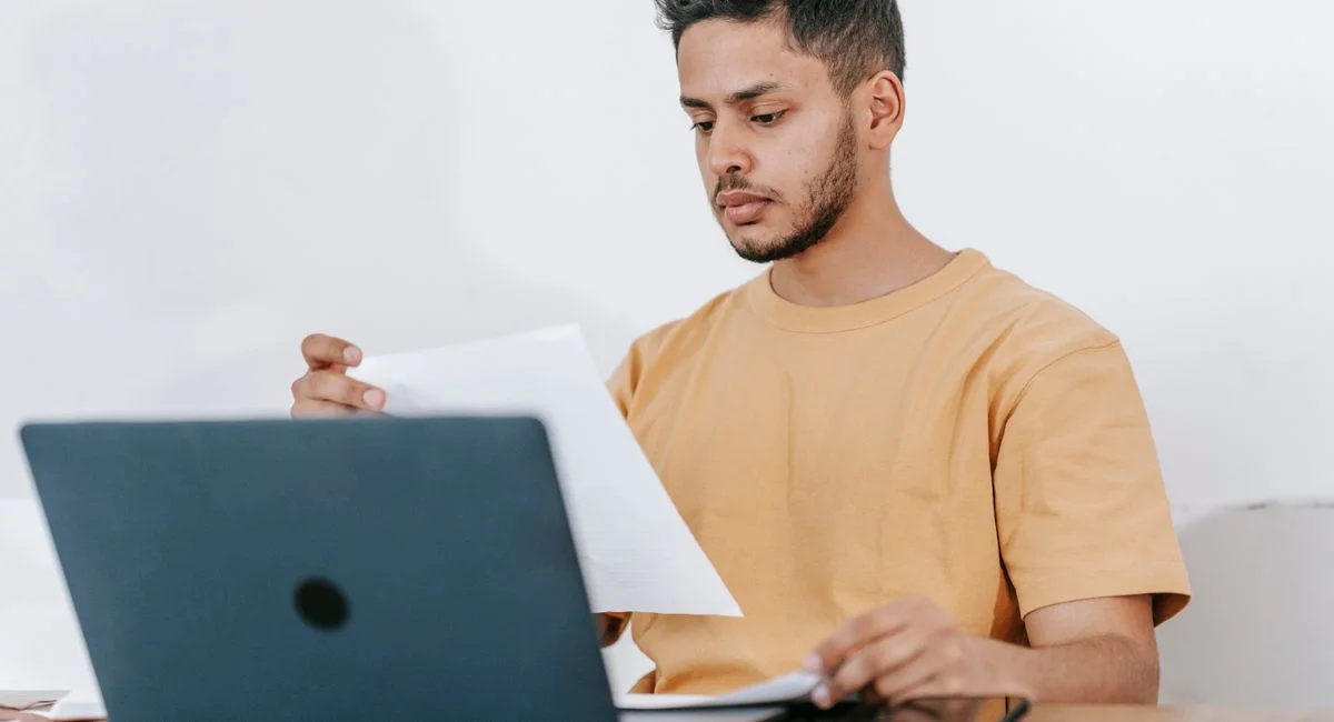 Young bearded Hispanic male university student reading information on paper while studying online via laptop