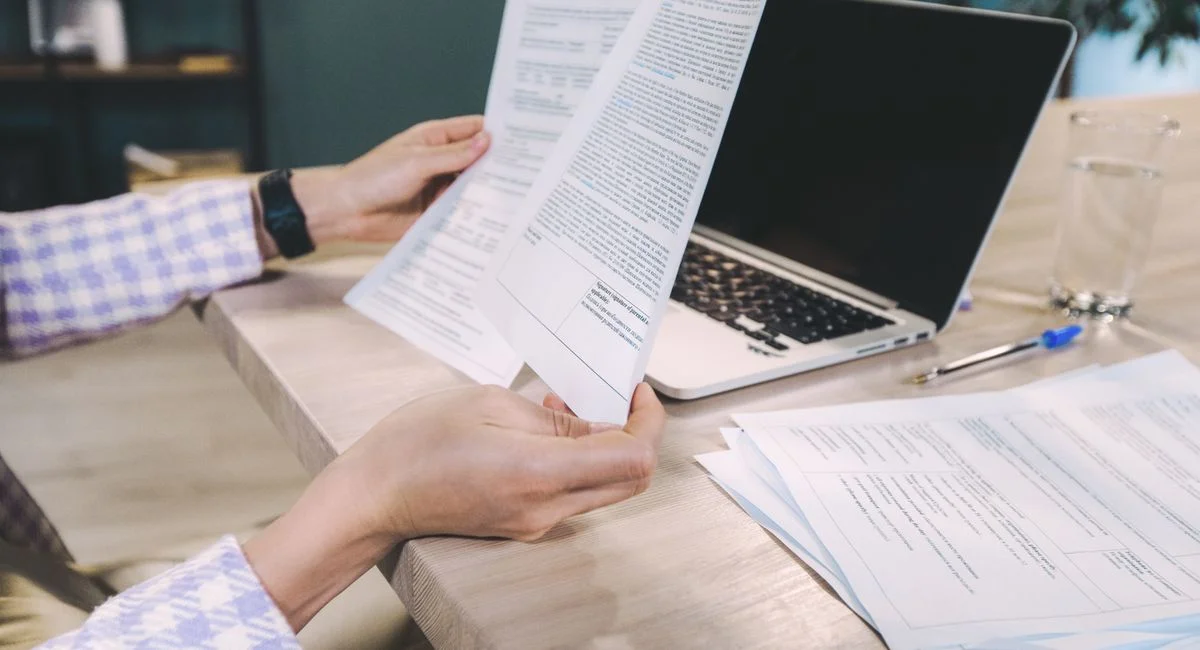 Close-up of hands examining printed documents next to laptop on office desk.