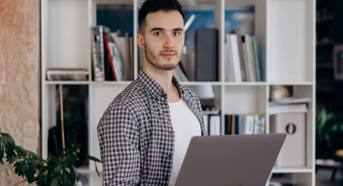 Man holding laptop in modern office setting with shelves. Professional and determined expression.