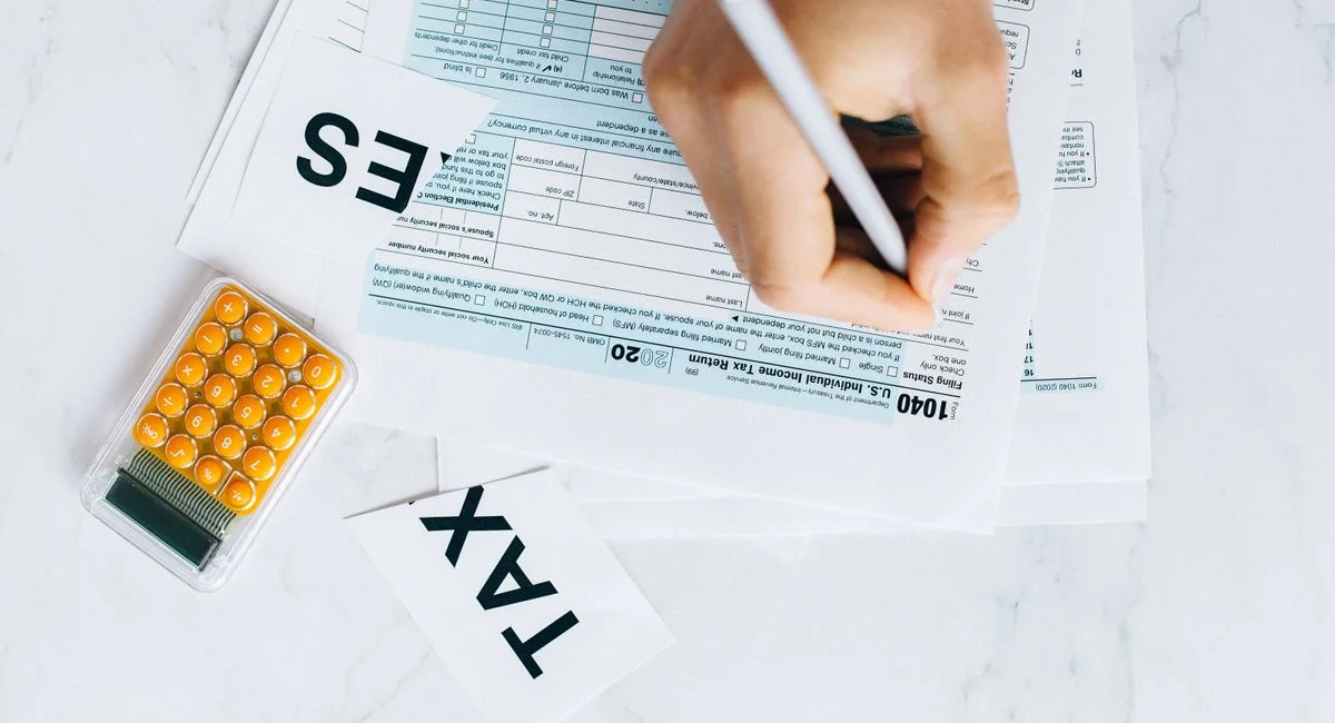 Close-up of hand writing on tax form with calculator nearby on white surface.
