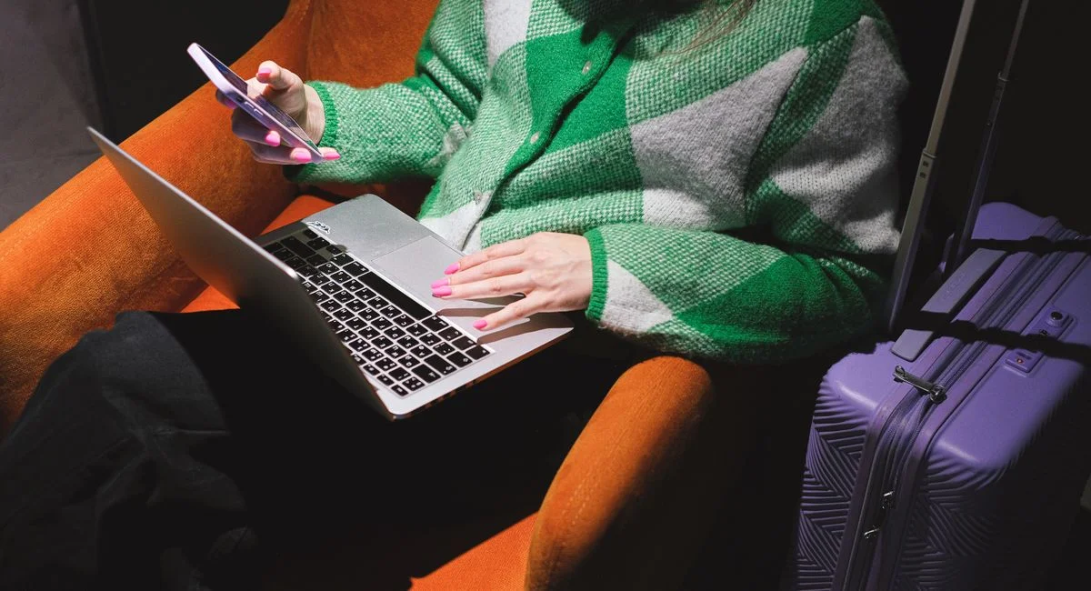 Woman in a knitted sweater working remotely on laptop with a smartphone, sitting in an armchair.