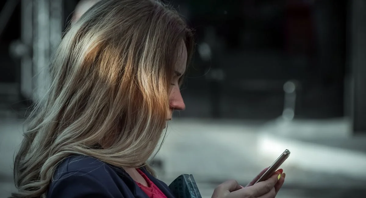 Side profile of a young woman texting on her smartphone in a shaded outdoor area.