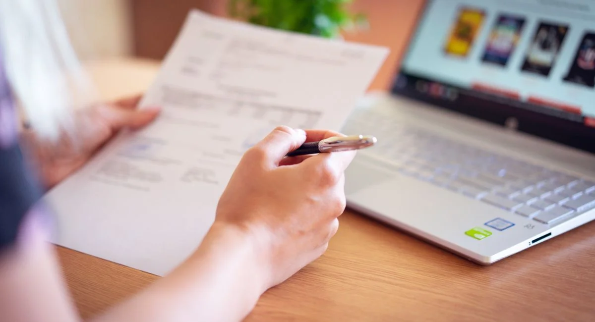 Close-up of a hand holding a pen and paper near a laptop, suggesting online shopping or document review.