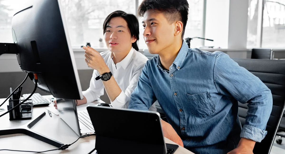 Two men collaborating on a project at a desktop computer in a bright office setting.