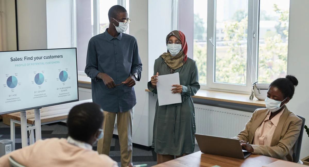 Office meeting with diverse colleagues wearing masks, presenting sales data.
