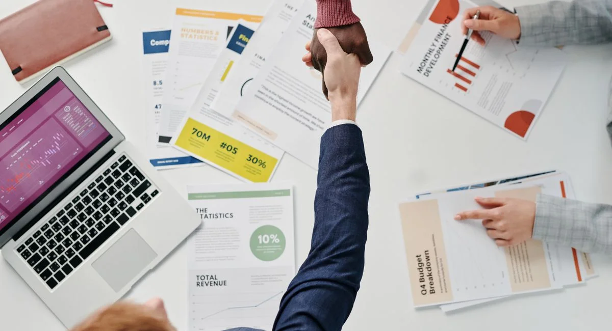 Business professionals engaging in a handshake over a desk filled with financial documents and a laptop.