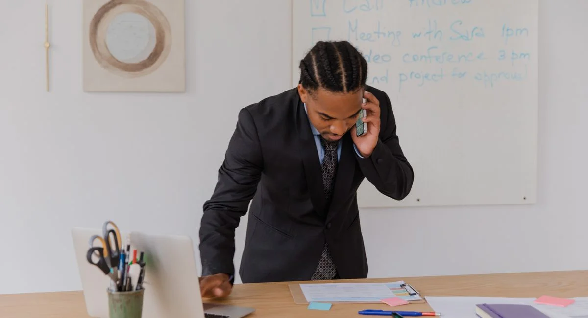 Professional businessman multitasking with phone and laptop at office desk, surrounded by documents.
