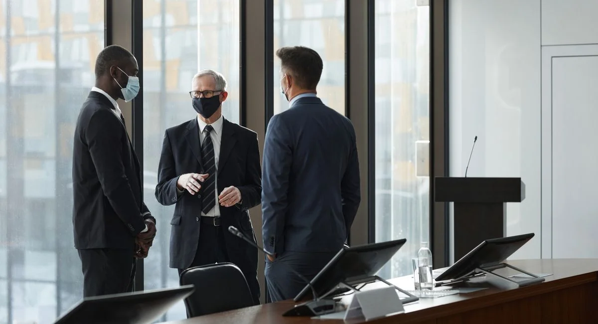 Three businessmen in suits and masks discussing in a modern conference room.
