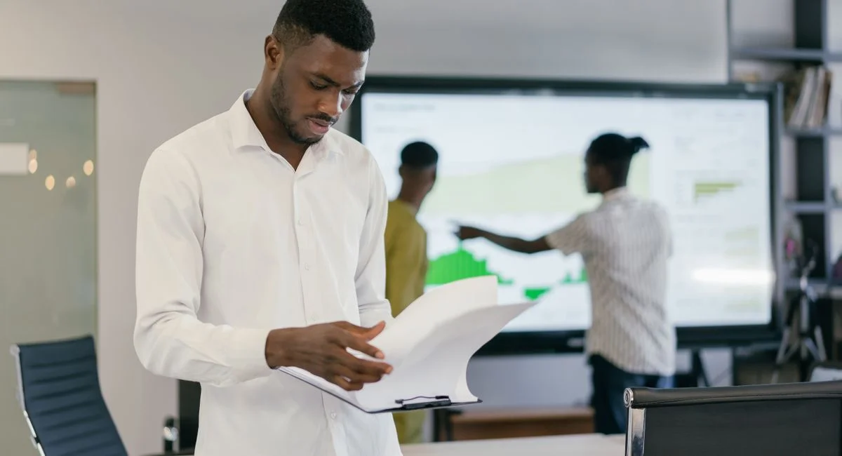 Man in office reading documents with colleagues in the background, strategizing business approaches.